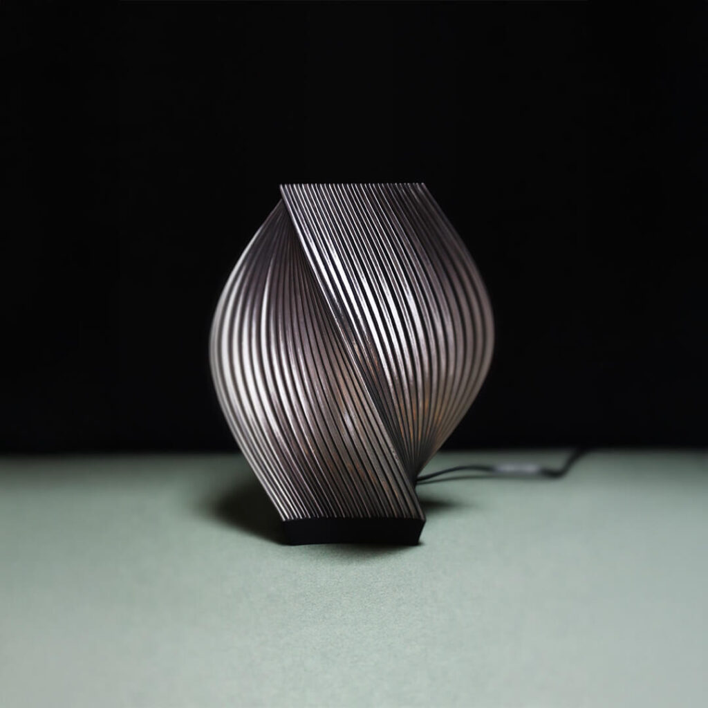 Lampada da tavolo MERAK di Cosmo Design, stampata in 3D con struttura elicoidale e luce diffusa, fotografata su sfondo scuro.